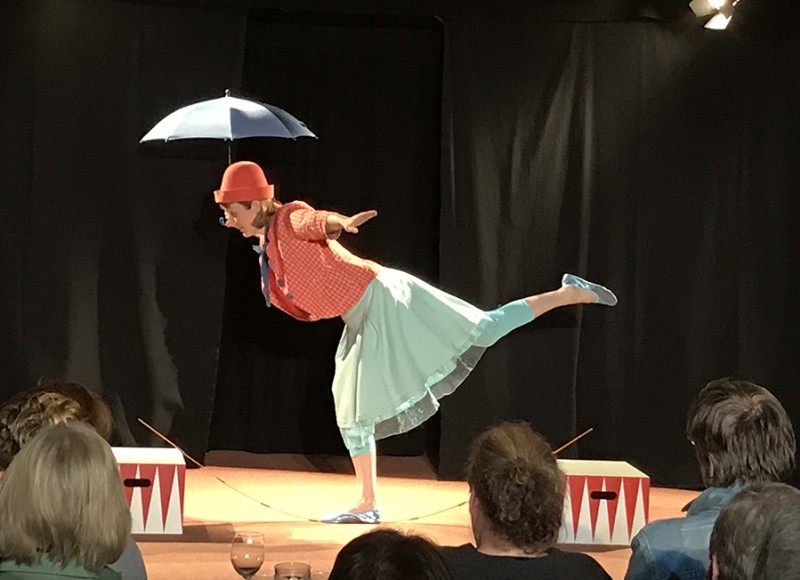 Clownin mit Schirm ist auf der Mitte der Bühne und balanciert auf einem Bein.